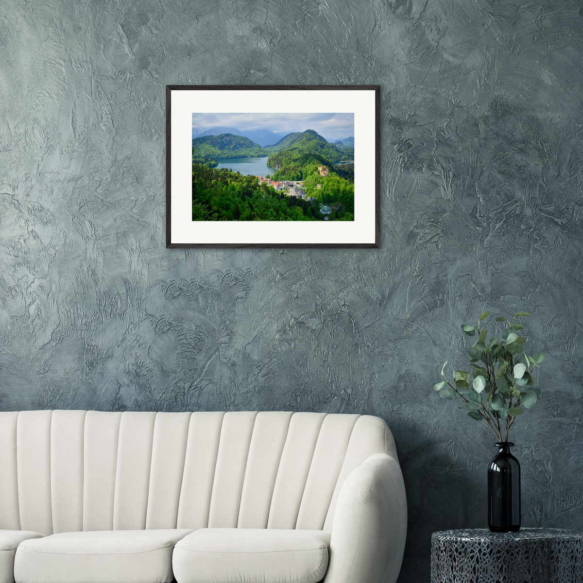 „Das Tal der Schlösser“ – gerahmte Fine-Art-Fotografie von Stanislav Shnaider in einem modernen Interieur mit grauer Strukturwand und weißem Sofa. Landschaft der Bayerischen Alpen mit Seen, bewaldeten Bergen und historischen Schlössern. Limitierte Fotokunst im Format 60 × 80 cm.