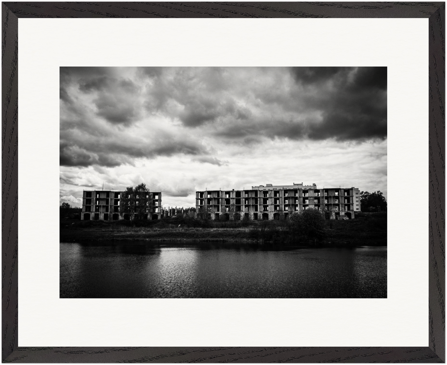 Gerahmte Schwarz-Weiß-Fotografie verlassener Wohngebäude am Ufer eines Sees unter schwerem Himmel.