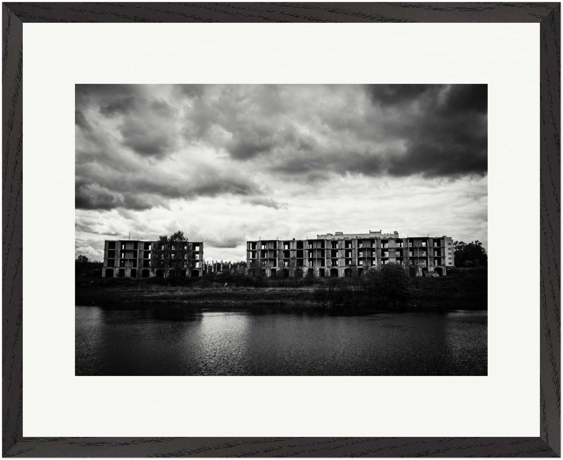 Gerahmte Schwarz-Weiß-Fotografie verlassener Wohngebäude am Ufer eines Sees unter schwerem Himmel.
