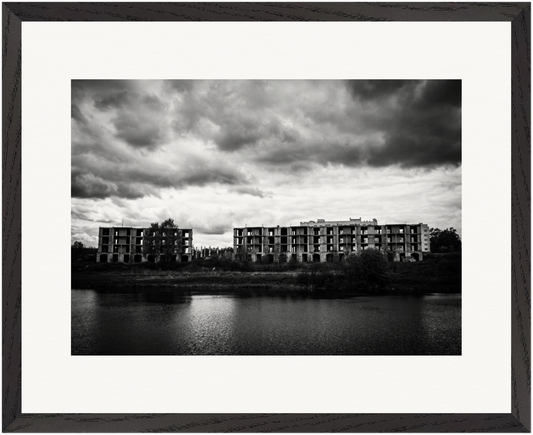 Gerahmte Schwarz-Weiß-Fotografie verlassener Wohngebäude am Ufer eines Sees unter schwerem Himmel.