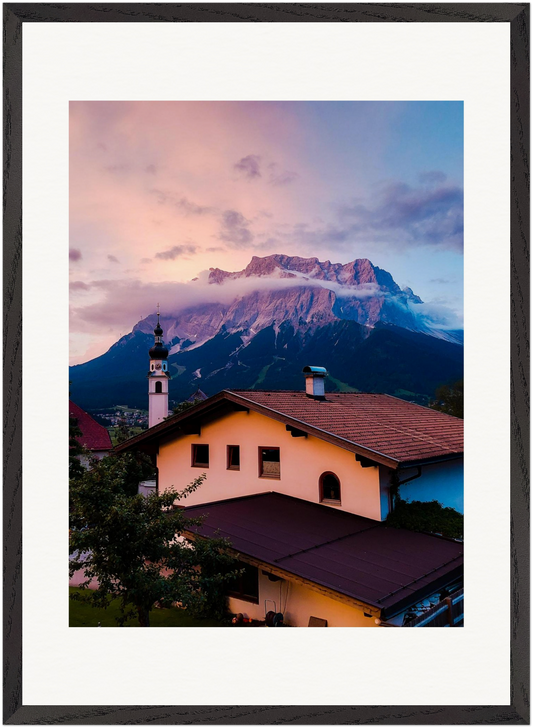Fine-Art-Fotografie der Zugspitze, aufgenommen von der österreichischen Seite in Tirol. Im Vordergrund ein traditionelles Tiroler Haus und eine Kirche, darüber die Alpen und ein eindrucksvoller Himmel. Originale Fine-Art-Fotografie von Stanislav Shnaider.